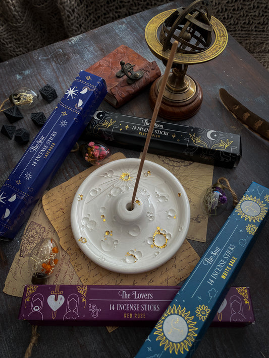 Assortimento di scatole di incensi colorate con temi astrologici, tra cui "The Moon" e "The Lovers", disposte accanto a un portaincenso ceramico bianco con dettagli dorati. Completano la scena oggetti mistici, tra cui un antico strumento astronomico, dadi runici e una bustina di fiori secchi, su un tavolo di legno con sottofondo di carta manoscritta, evocando un'atmosfera di antica saggezza e rituali calmanti.