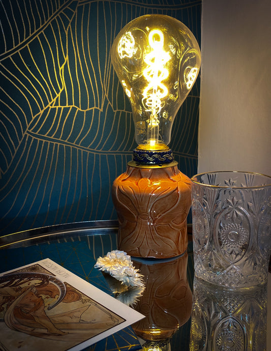 Lampada in ceramica arancione dal design Liberty con luce accesa, accanto a bicchiere di cristallo e carta tarocchi, che evoca un'atmosfera vintage.